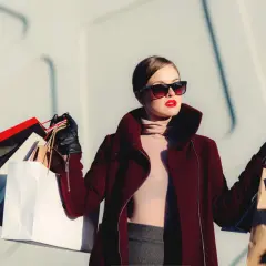 En kvinna med solglasögon och röda läppar bär fina höstjackor och shoppingkassar.
