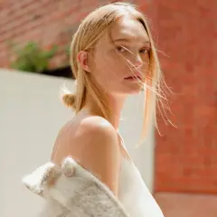 Gemma Ward med blont hår och en vit klänning, med en ullig jacka över axlarna, står framför en tegelvägg.