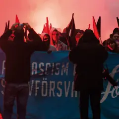 En grupp demonstranter med facklor och banderollen "ASCISM FÖRSVANN" och "FREE Joel". De agerar som en anti-hjälte, som kämpar mot orättvisor.