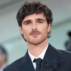 Jacob Elordi style: en ung man med mörkt hår och mustasch bär en svart kostym med en fjärilsbrosch.
