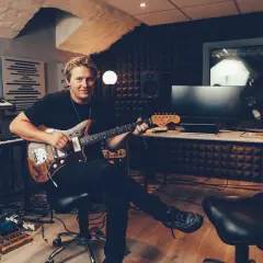 John Engelbert sitter i en inspelningsstudio med en gitarr i famnen, omgiven av musikinstrument och teknisk utrustning.