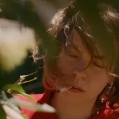 En ung kvinna med slutna ögon och en cigarett i munnen, omgiven av grönska. Scenen påminner om stämningen i sorgliga filmer.