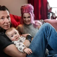 En familj med ett etype barn myser i soffan. Pappan håller barnet, mamman med rosa hår ler.