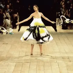 Shalom Harlow ung i en vit klänning med svart och gul spraymålning, omgiven av robotarmar på en catwalk.
