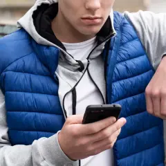 Ung kille med blå dunjacka och grå hoodie kollar på sin mobil. Kanske undrar han vad betyder simp?