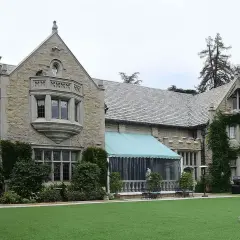 En person går över den gröna gräsmattan framför Playboy Mansion, en ikonisk byggnad med stenfasad och tak av skiffer.
