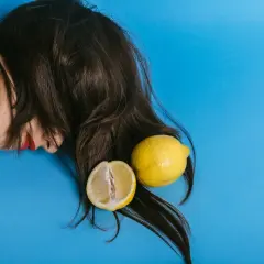 En person med mörkt hår och röda läppar ligger på en blå bakgrund med en hel och en halv citron. Kanske en idé för att bleka håret med citron?