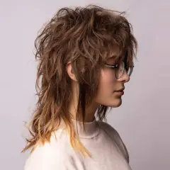 En ung kvinna med glasögon bär en shaggy mullet frisyr med rufsigt, brunt hår som bleknar till blont i topparna.
