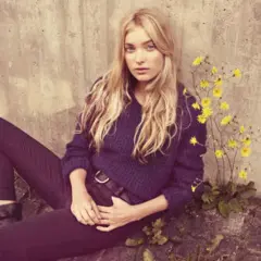 Elsa Hosk, med sitt blonda hår och blå ögon, sitter avslappnat mot en betongvägg, omgiven av gula blommor.