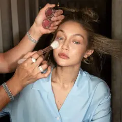En makeupartist applicerar puder med en borste på en modell. Två små burkar med rouge syns i handen. En perfekt finish för baking makeup.