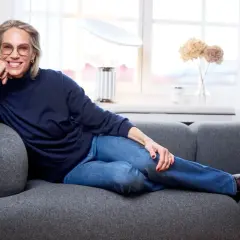 Tyra Wiklund, med ett leende, vilar på en grå soffa i jeans och mörkblå tröja.