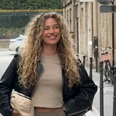 En kvinna med vackert lockigt hår ler och tittar bort. Hon bär en svart jacka och en beige tröja.