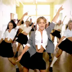 En grupp dansare, med fokus på en blond kvinna i mitten, utför en koreografi i en skolkorridor. Deras **blonde halloween costumes** är inspirerade av skoluniformer.
