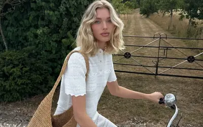 En kvinna med blont hår på en cykel, klädd i ett vitt sommar set och med en stor stråväska.