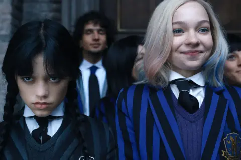 Skådespelare Wednesday Addams med flätor och en allvarlig min, bredvid en leende elev med blont hår och blåa inslag.