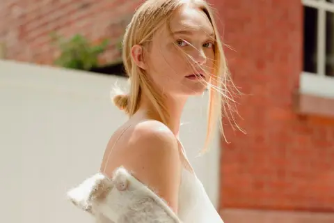 Gemma Ward med blont hår och en vit klänning, med en ullig jacka över axlarna, står framför en tegelvägg.