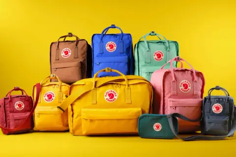 En samling färgglada Fjällräven Kånken-ryggsäckar i olika fjällräven ryggsäck storlek, från små till stora, mot en gul bakgrund.