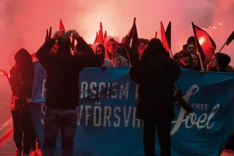En grupp demonstranter med facklor och banderollen "ASCISM FÖRSVANN" och "FREE Joel". De agerar som en anti-hjälte, som kämpar mot orättvisor.