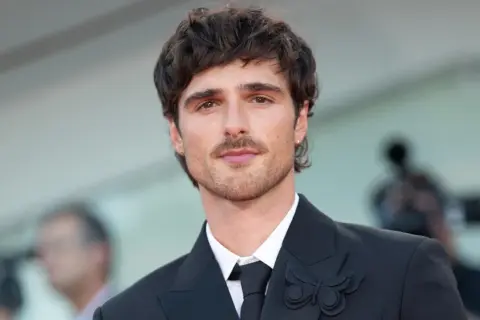 Jacob Elordi style: en ung man med mörkt hår och mustasch bär en svart kostym med en fjärilsbrosch.