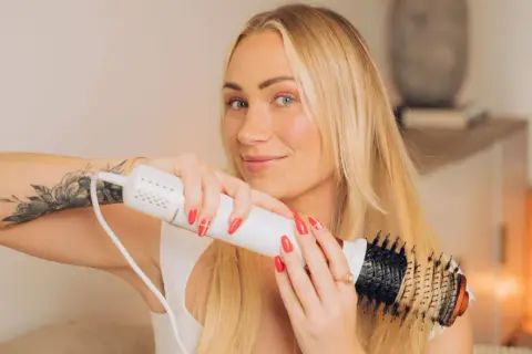 Blond kvinna med tatuering på armen stylar sitt hår med en varmluftsborste för perfekt blow out hår.