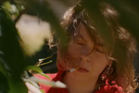 En ung kvinna med slutna ögon och en cigarett i munnen, omgiven av grönska. Scenen påminner om stämningen i sorgliga filmer.