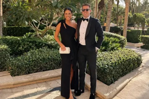 Ett par klädda för black tie dresscode, hon i en elegant svart klänning, han i smoking. De poserar glatt utomhus bland grönska.