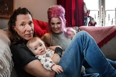 En familj med ett etype barn myser i soffan. Pappan håller barnet, mamman med rosa hår ler.