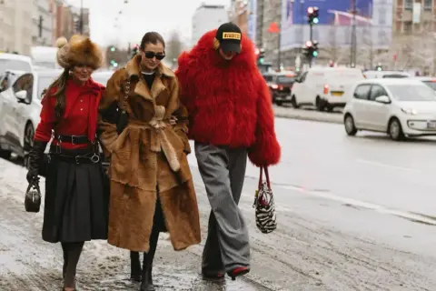 Tre kvinnor i trendiga ytterkläder, en med röd pälsjacka, visar upp **copenhagen street style** på en snöig gata.