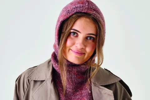 Ung kvinna med blå ögon bär en stickad balaclava i rosa och lila nyanser, med en beige trenchcoat.
