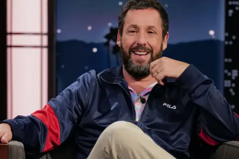 Adam Sandler skrattar glatt i en blå Fila-jacka, redo för en rolig intervju.