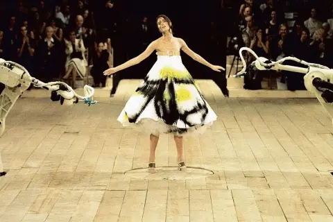 Shalom Harlow ung i en vit klänning med svart och gul spraymålning, omgiven av robotarmar på en catwalk.