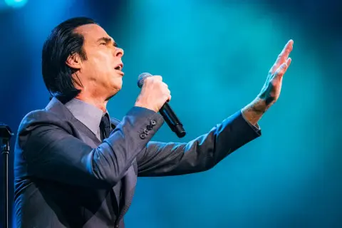 Nick Cave sjunger passionerat med mikrofonen i handen under en nick cave tour. Hans arm är utsträckt mot en blå bakgrund.