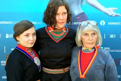 Tre kvinnor i traditionella samiska dräkter poserar framför en blå bakgrund med logotyper. De representerar en stark berättelse, likt den i feministiska filmer.