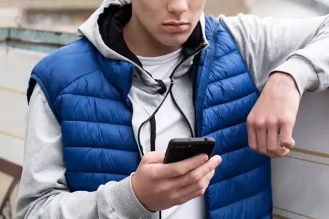 Ung kille med blå dunjacka och grå hoodie kollar på sin mobil. Kanske undrar han vad betyder simp?
