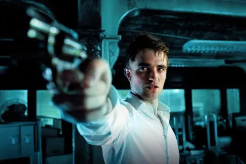 Robert Pattinson i en vit skjorta siktar med en pistol i en mörk, mystisk scen, typisk för hans filmer.
