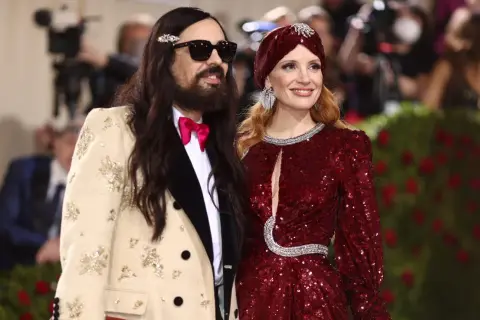 Alessandro Michele och Jessica Chastain poserar glatt. Han bär en beige kavaj med broderier och en rosa fluga.
