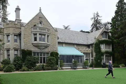 En person går över den gröna gräsmattan framför Playboy Mansion, en ikonisk byggnad med stenfasad och tak av skiffer.