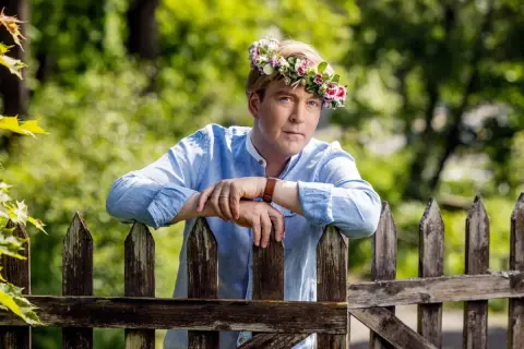 En man med blomsterkrans på huvudet lutar sig mot ett staket. En sommarprat recension som fångar essensen av svensk sommar.