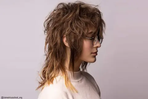 En ung kvinna med glasögon bär en shaggy mullet frisyr med rufsigt, brunt hår som bleknar till blont i topparna.