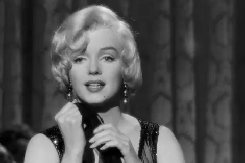 Marilyn Monroe i en ikonisk scen från en klassisk film. Hennes blonda lockar, pärlörhängen och glittriga klänning fångar blicken.
