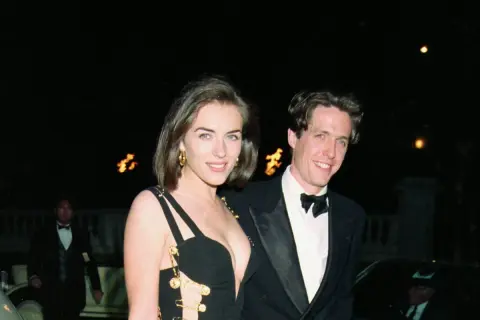 Elizabeth Hurley 90s i Hugh Grant ler mot kameran. Hon bär en ikonisk svart klänning med gulddetaljer.