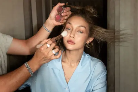 En makeupartist applicerar puder med en borste på en modell. Två små burkar med rouge syns i handen. En perfekt finish för baking makeup.