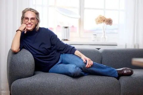Tyra Wiklund, med ett leende, vilar på en grå soffa i jeans och mörkblå tröja.