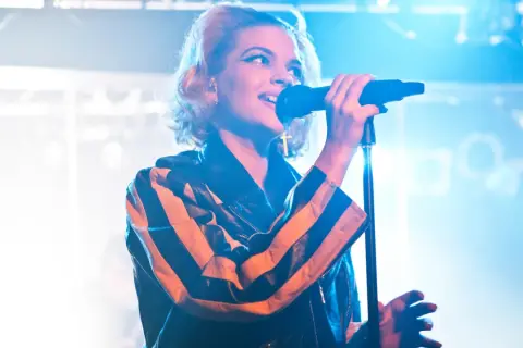En artist med tove styrke sjunger i mikrofonen, iklädd en läderjacka med orange och svarta ränder.
