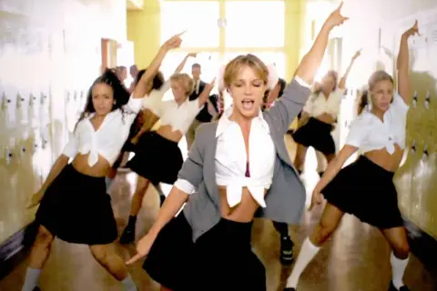 En grupp dansare, med fokus på en blond kvinna i mitten, utför en koreografi i en skolkorridor. Deras **blonde halloween costumes** är inspirerade av skoluniformer.