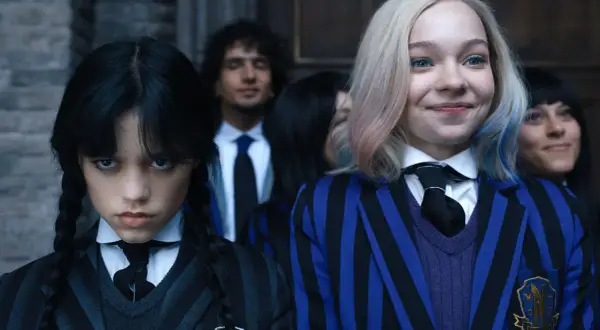 Skådespelare Wednesday Addams med flätor och en allvarlig min, bredvid en leende elev med blont hår och blåa inslag.