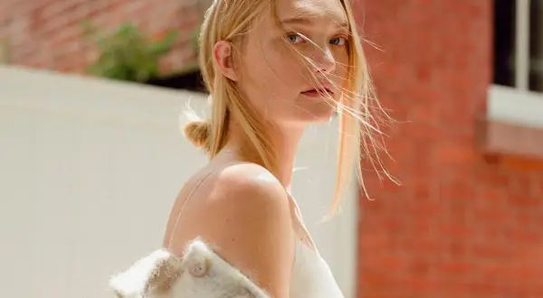 Gemma Ward med blont hår och en vit klänning, med en ullig jacka över axlarna, står framför en tegelvägg.