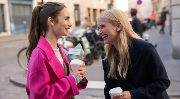 Camille och Emily i Paris, skrattandes med kaffe och blommor på en solig gata.