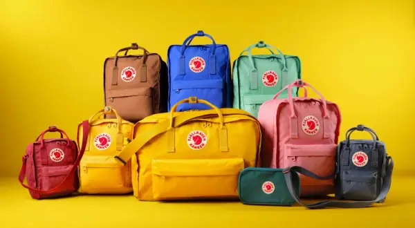 En samling färgglada Fjällräven Kånken-ryggsäckar i olika fjällräven ryggsäck storlek, från små till stora, mot en gul bakgrund.