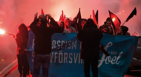 En grupp demonstranter med facklor och banderollen "ASCISM FÖRSVANN" och "FREE Joel". De agerar som en anti-hjälte, som kämpar mot orättvisor.